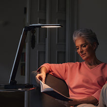 Load image into Gallery viewer, A user using the Elumentis Lamp on a side table to read a book.