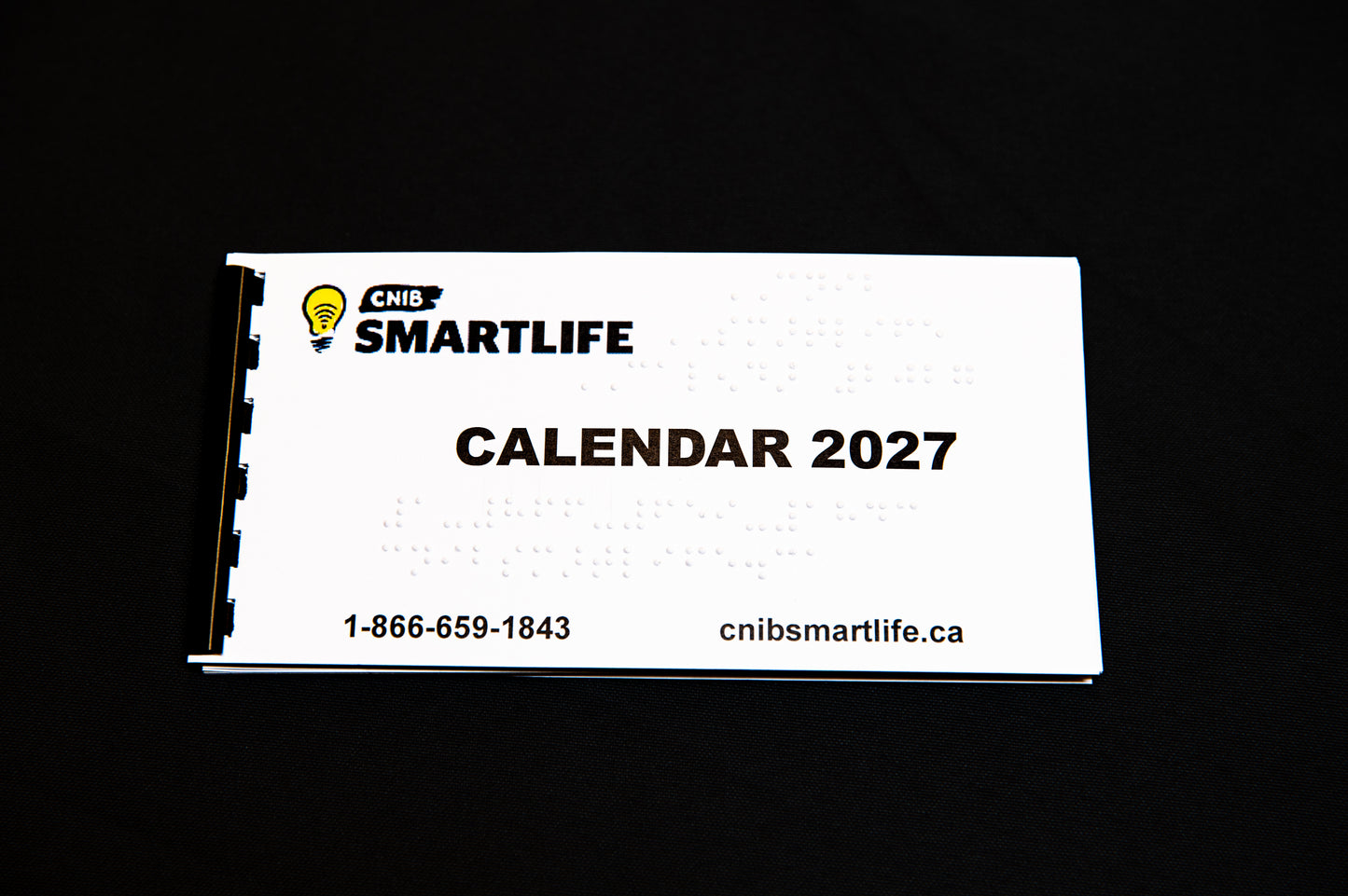 A white spiral-bound 2027 Braille calendar with black text and a black-and-white spiral binding edge, photographed on a solid bright black background.
