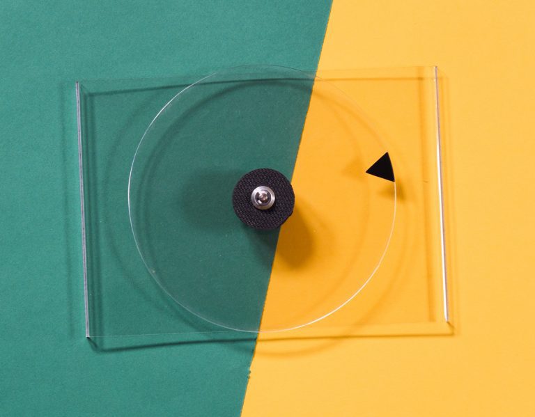 Clear plastic spinner with a black centre knob and pointer, displayed on a background divided into green and yellow sections.