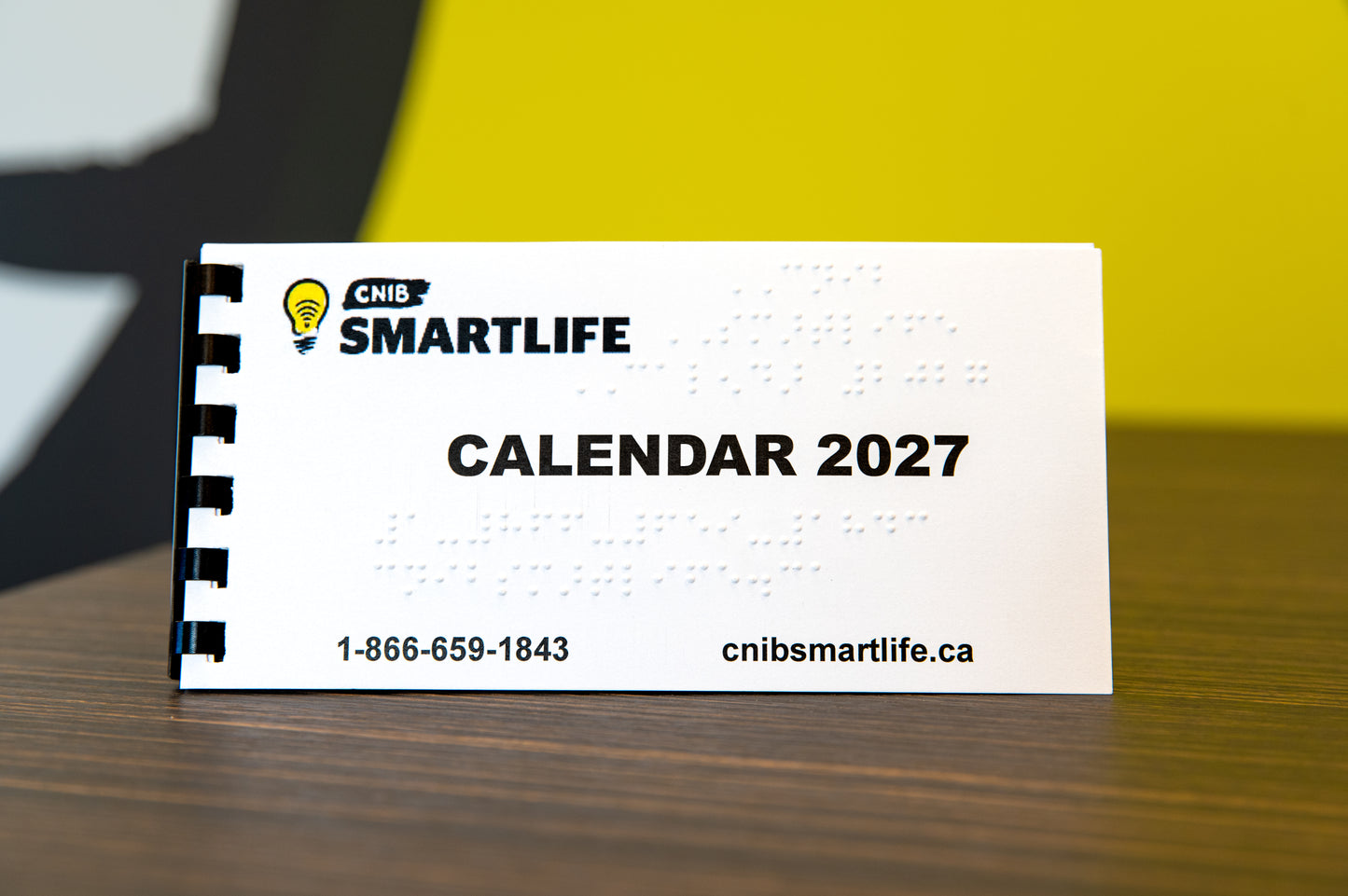 A white spiral-bound 2027 Braille calendar from CNIB Smartlife, featuring black text and Braille, displayed on a wooden table with a yellow background.
