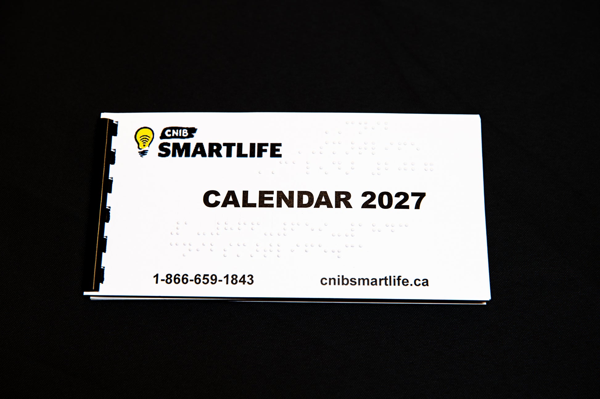 A white spiral-bound 2027 Braille calendar with black text and a black-and-white spiral binding edge, photographed on a solid bright black background. 