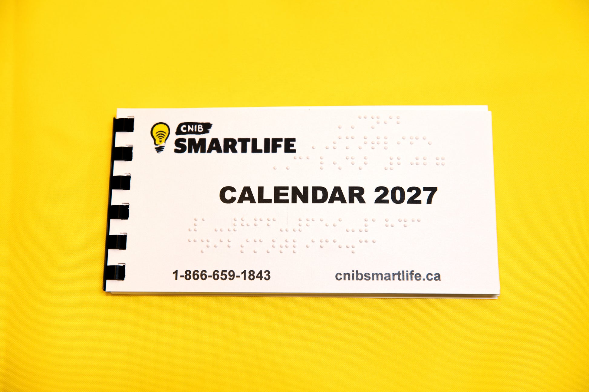 A white spiral-bound 2027  Braille calendar with black text and a black-and-white spiral binding edge, photographed on a solid bright yellow background. 