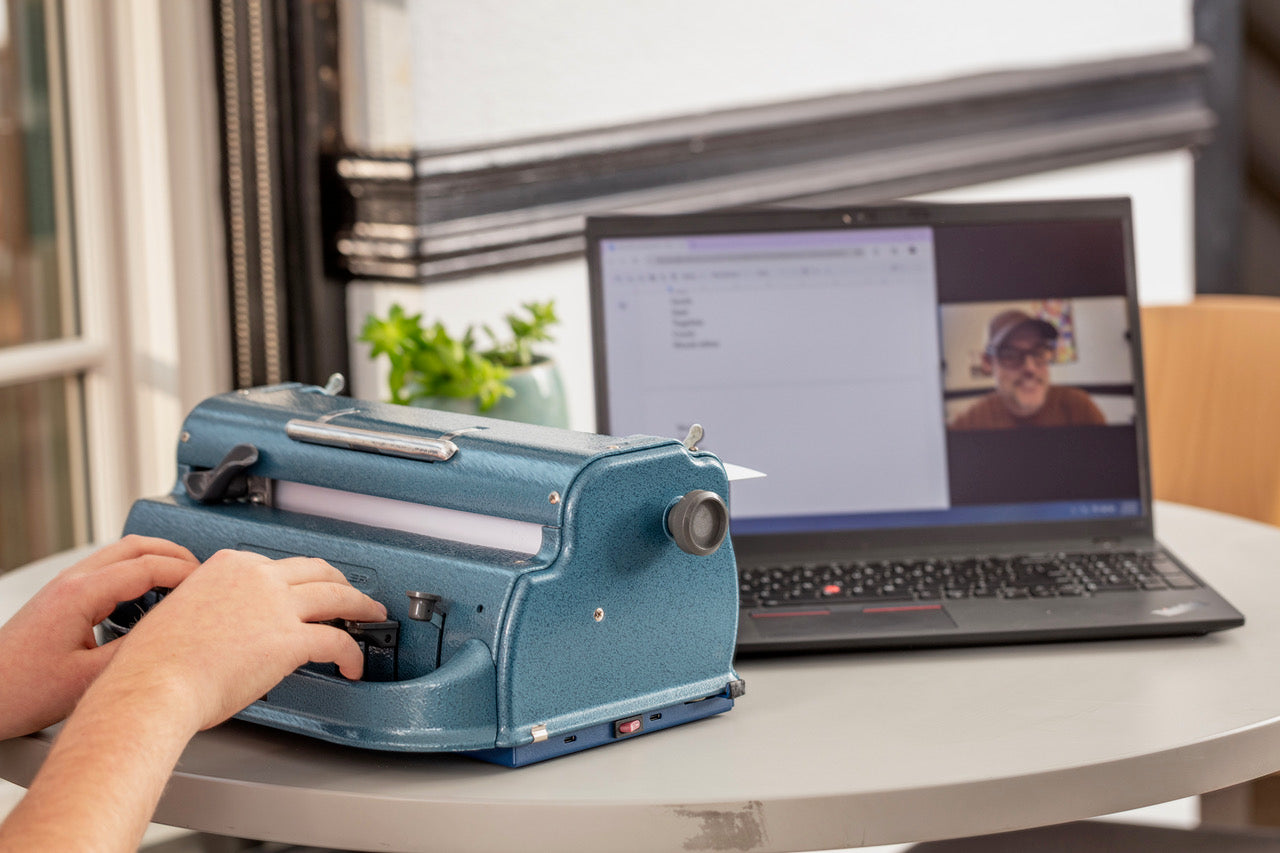 Person using a Perkins Blue Brallier with the Perkins Bloom attached with a laptop displaying a video call in the background.