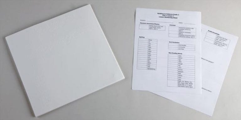 A closed white binder beside printed lesson-monitoring sheets containing tables and assessment information.