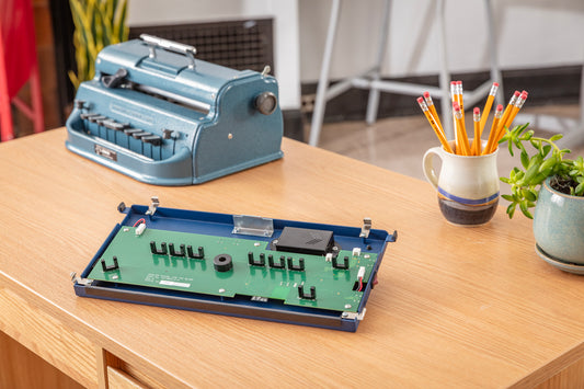 Displayed is a Perkins Blue Braille Bloom device with a Perkins Blue Brailler in the background on a brown desk 