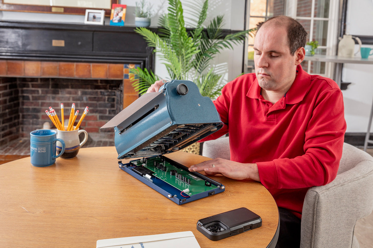 A Man with the Perkins Blue  Braille Bloom installing onto a Perkins Brailler demonstrating how the device connects underneath at a desk with a home office setting.