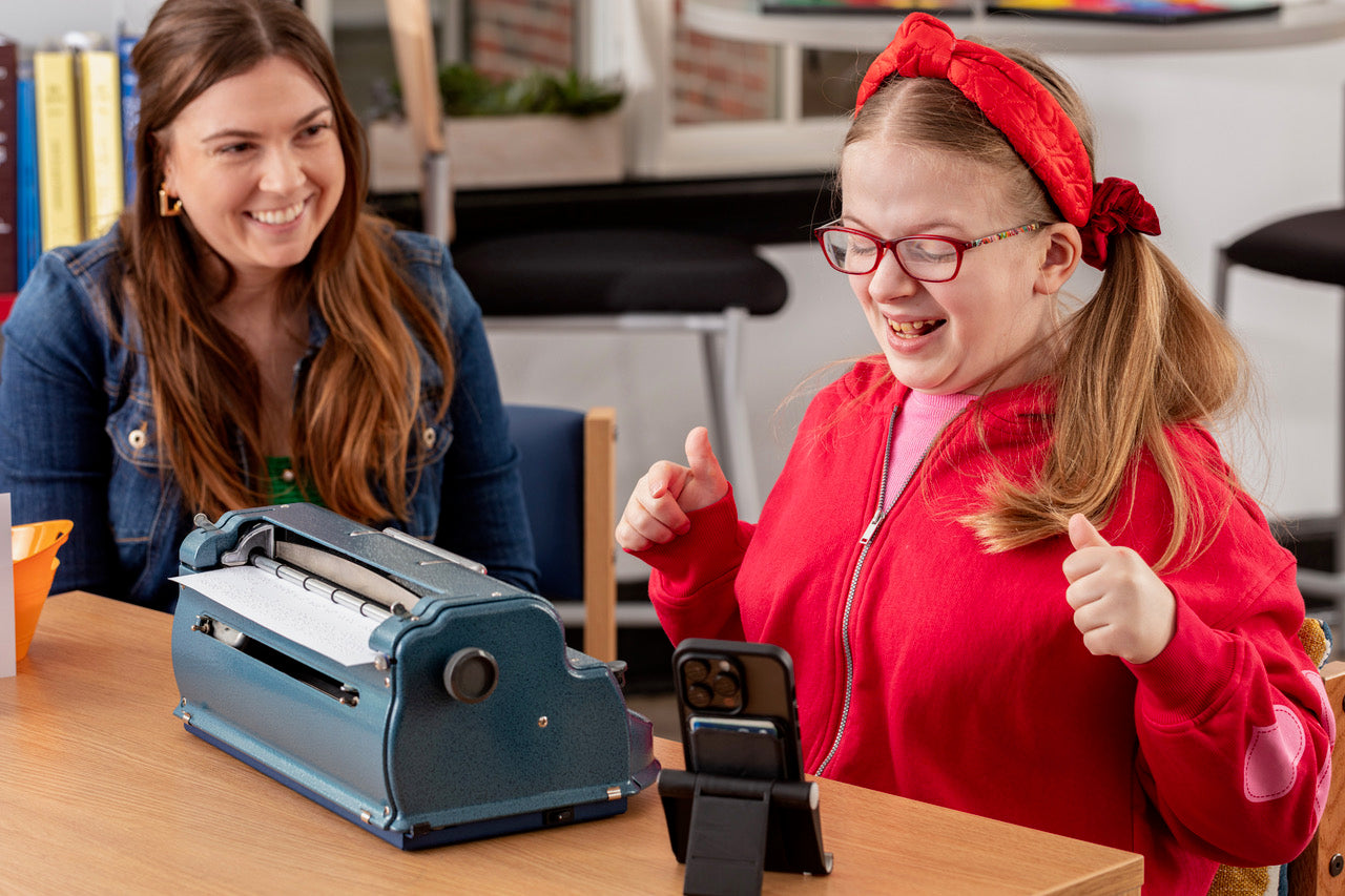 Student using a Perkins blue Brailler with Braille Bloom while a teacher views the text output on a screen