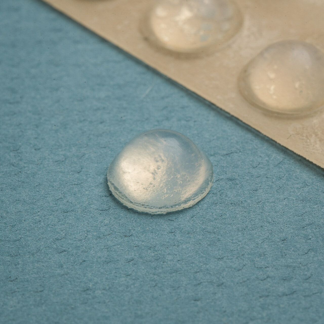 A close-up of a single clear, round tactile bump dot used for marking appliances, with more dots visible in the background. on a blue surface.